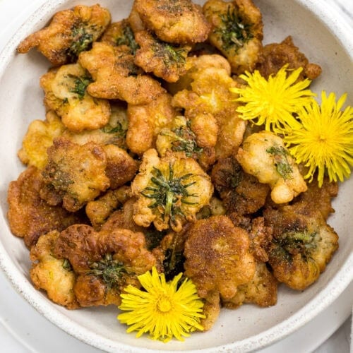 A bowl of golden brown, battered and fried dandelion greens, garnished with yellow dandelion flowers, resembling irresistible Dandelion Donuts.