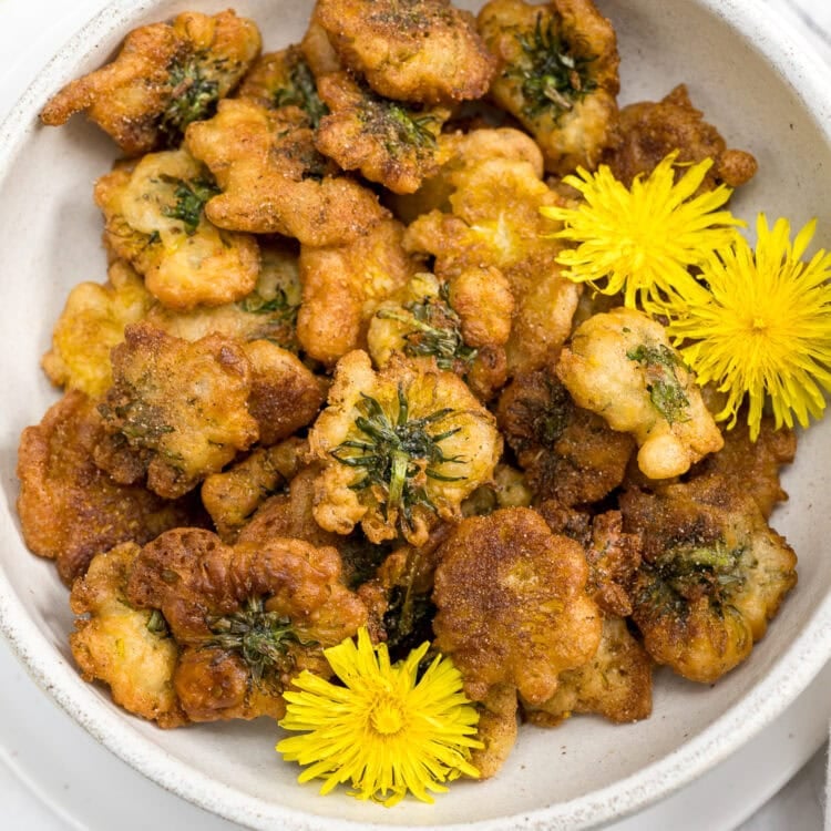 A bowl of golden brown, battered and fried dandelion greens, garnished with yellow dandelion flowers, resembling irresistible Dandelion Donuts.