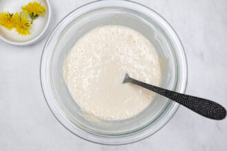 A glass bowl filled with batter and a spoon, placed on a white surface; a small bowl with dandelion flowers sits nearby, ready to be mixed in for homemade Dandelion Donuts.