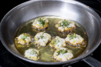 Seven stuffed squash blossoms are frying in hot oil in a stainless steel pan, their golden edges crisping up like Dandelion Donuts.