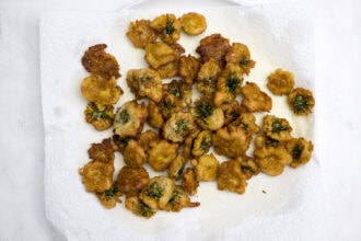 A batch of golden-brown, dandelion-infused fried food pieces with herbs rests on a white paper towel for draining oil.