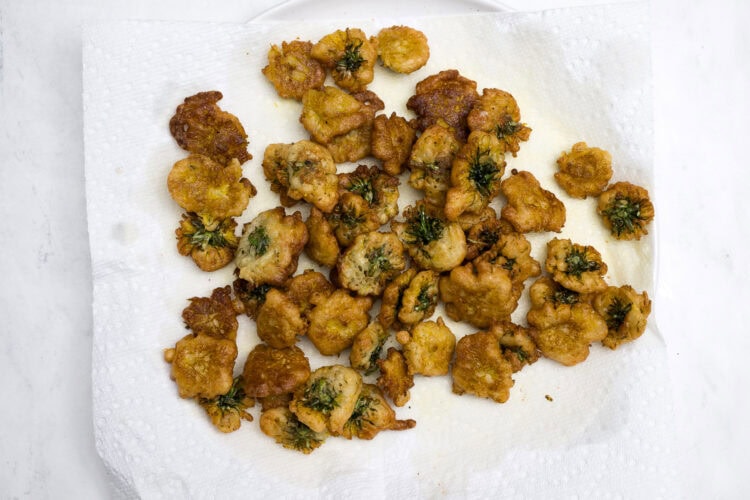 A batch of golden-brown, dandelion-infused fried food pieces with herbs rests on a white paper towel for draining oil.
