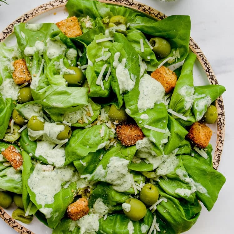 A plate of green salad with lettuce, green olives, croutons, shredded cheese, and herb Green Goddess Dressing. A glass jar of dressing and parsley are nearby on the white surface.