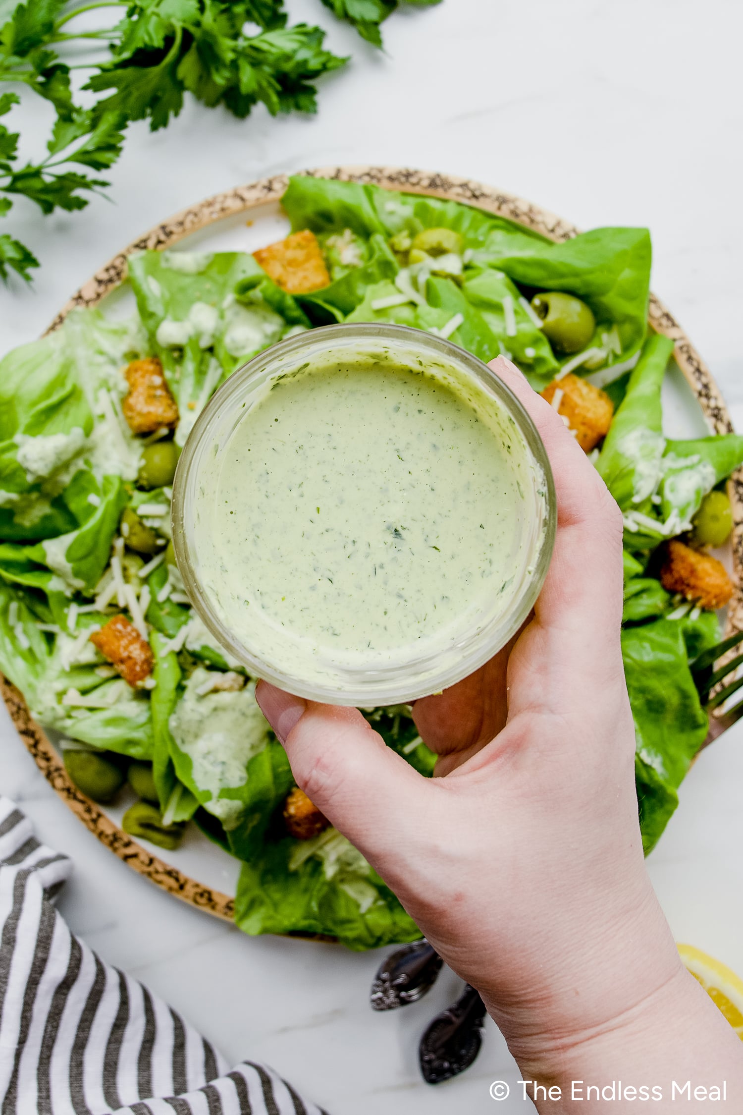 A hand holds a jar of creamy Green Goddess Dressing above a plate of green salad with lettuce, croutons, olives, and grated cheese.