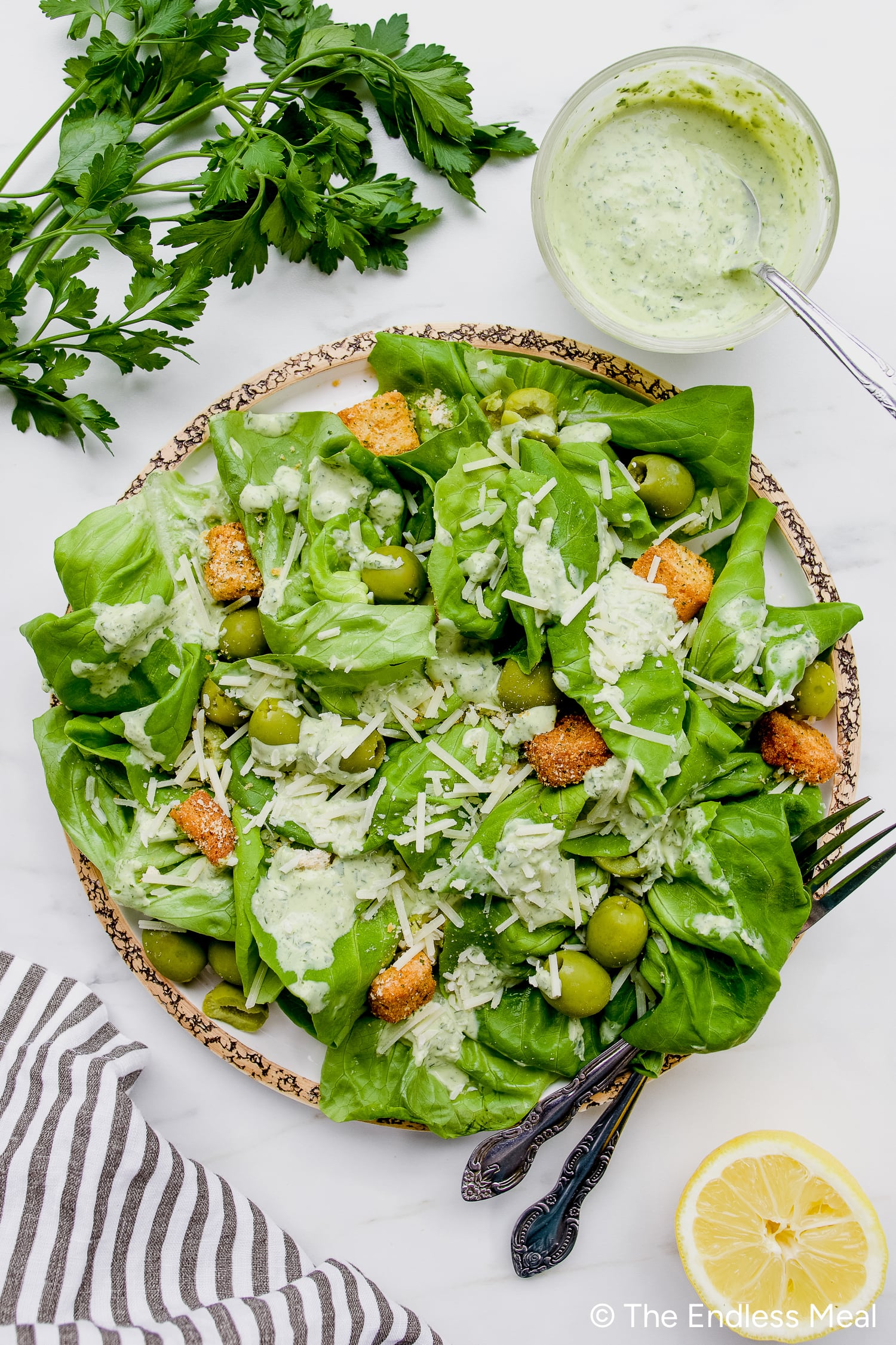 A plate of leafy green salad with green olives, croutons, shredded cheese, and creamy Green Goddess Dressing sits next to a bowl of dressing, parsley, lemon slice, and a striped napkin.