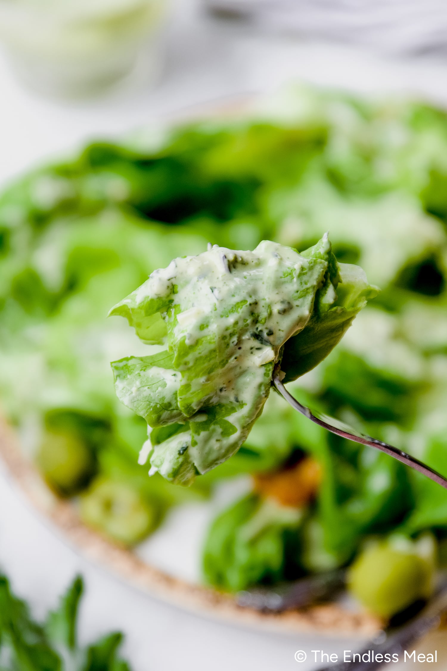 A fork holds a bite of green lettuce topped with creamy Green Goddess Dressing, with more salad visible on a plate in the blurred background.