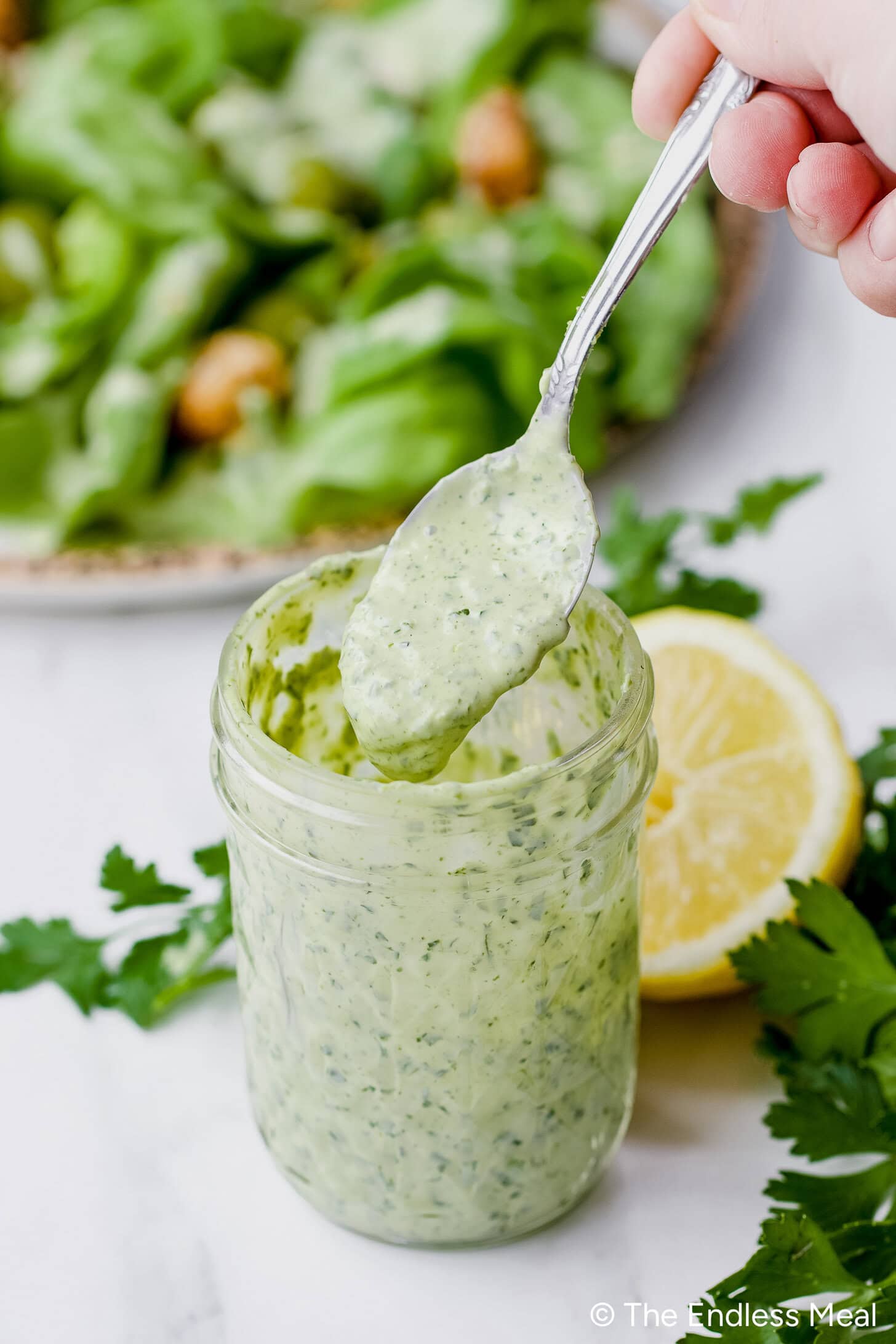 A spoon is lifting creamy Green Goddess Dressing from a glass jar, with parsley, lemon, and a salad in the background.