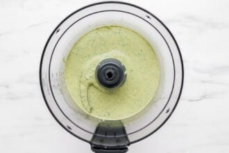 A food processor filled with a smooth, pale green Green Goddess Dressing is viewed from above on a white marble surface.