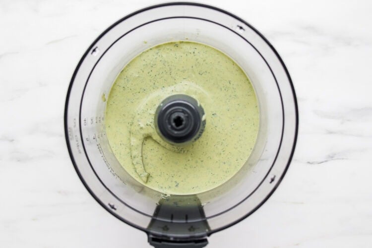 A food processor filled with a smooth, pale green Green Goddess Dressing is viewed from above on a white marble surface.