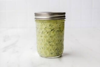 A glass mason jar filled with vibrant Green Goddess Dressing, sealed with a metal lid, sits on a white marble surface against a tiled background.