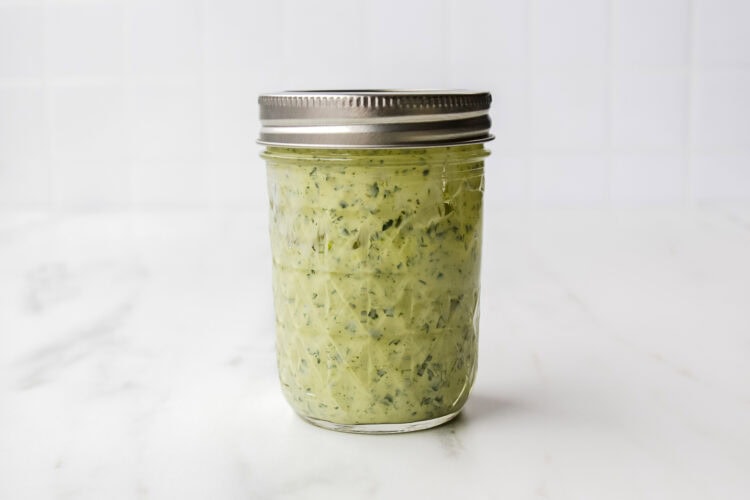A glass mason jar filled with vibrant Green Goddess Dressing, sealed with a metal lid, sits on a white marble surface against a tiled background.