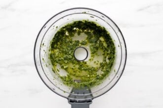 Overhead view of a food processor containing a vibrant Green Goddess Dressing herb mixture with oil on a white marble surface.
