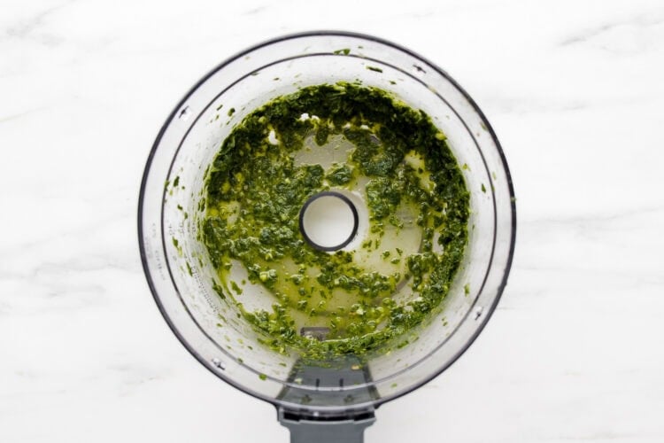 Overhead view of a food processor containing a vibrant Green Goddess Dressing herb mixture with oil on a white marble surface.