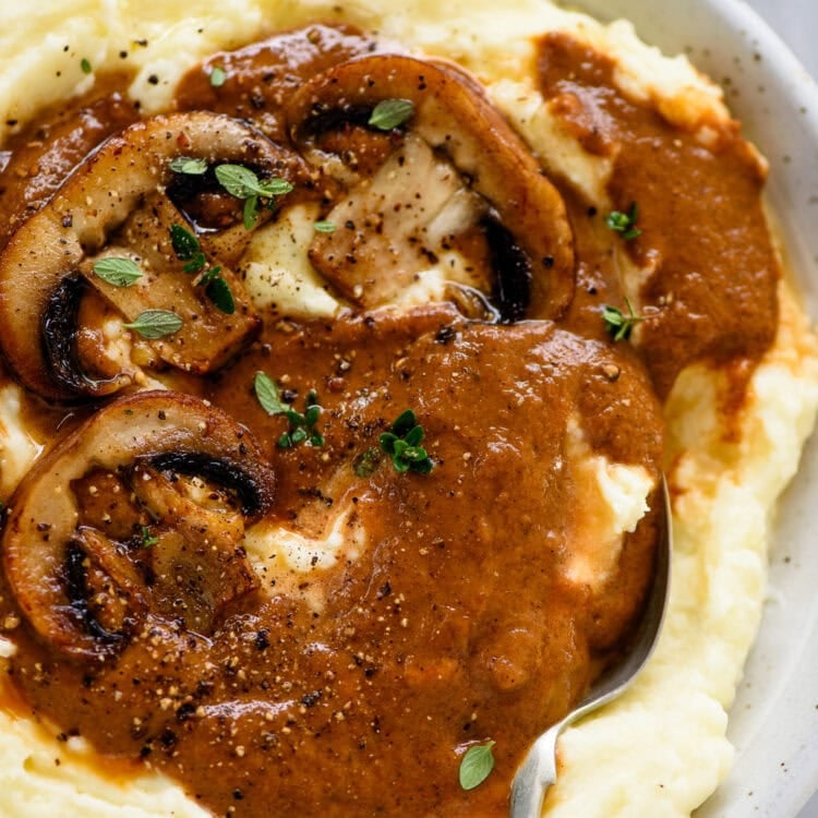 A bowl of mashed potatoes topped with savory mushroom gravy, garnished with fresh herbs and cracked black pepper.