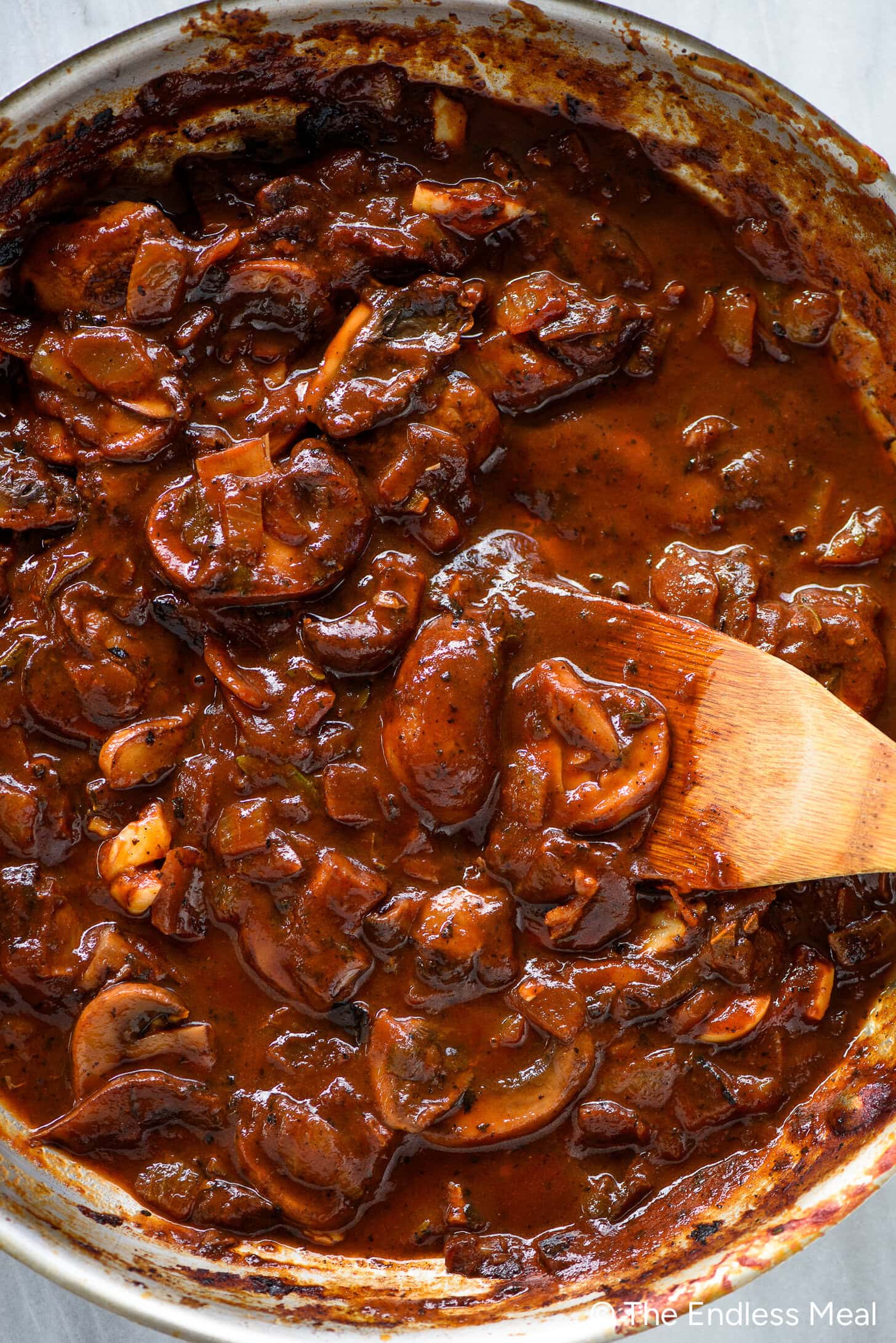A close-up of a skillet filled with mushroom and onion stew in a rich brown mushroom gravy, being stirred with a wooden spoon.