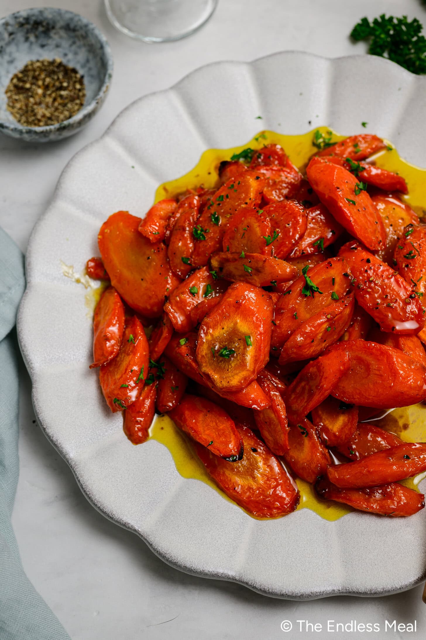 A plate of roasted Honey Garlic Carrots garnished with chopped herbs and drizzled with olive oil, set on a light surface next to a small bowl of pepper.