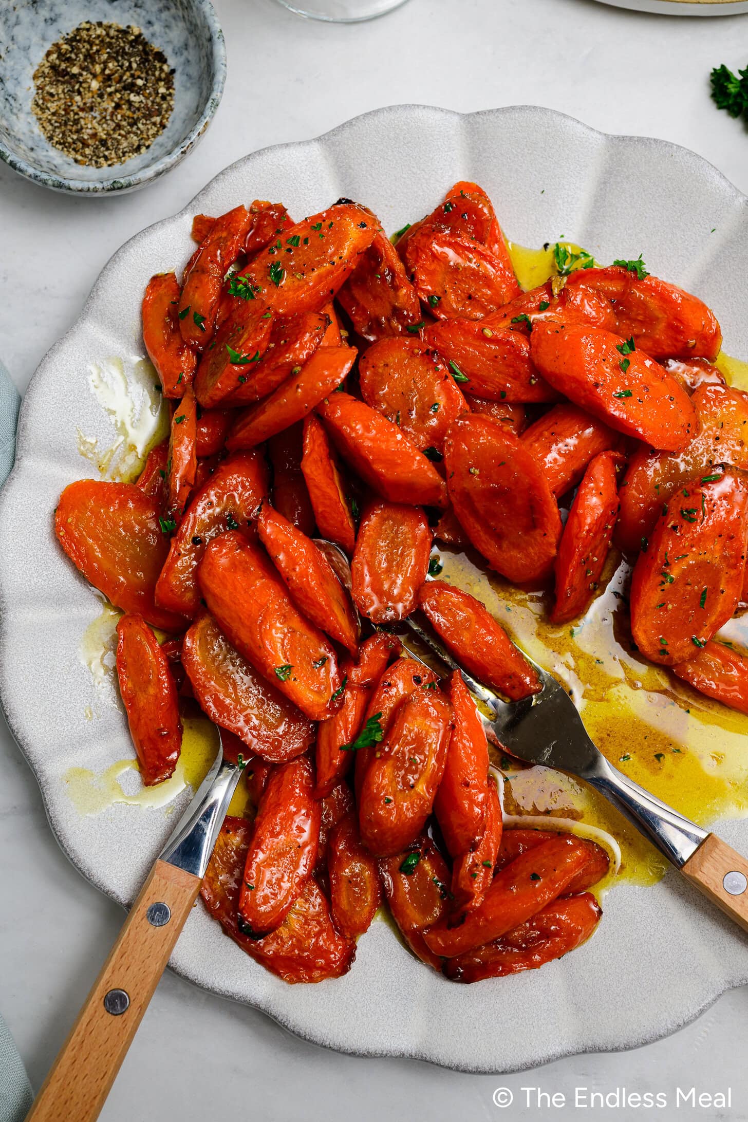 A white plate with sliced, roasted Brown Butter Carrots garnished with herbs and drizzled with olive oil, served with wooden-handled utensils.