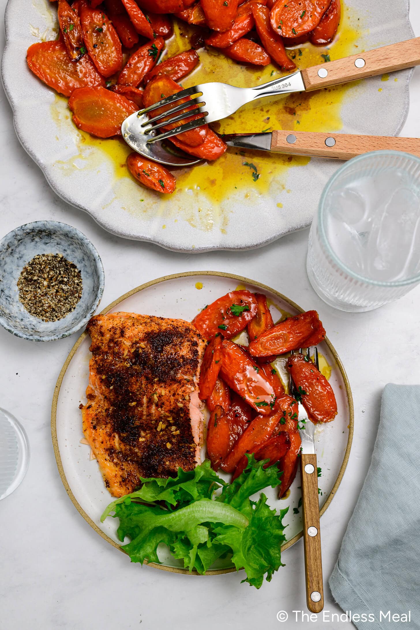 A plate with a seasoned salmon fillet, brown butter carrots, and lettuce. Nearby are a fork, a knife, a bowl of pepper, an empty glass, and a partially served dish of carrots.