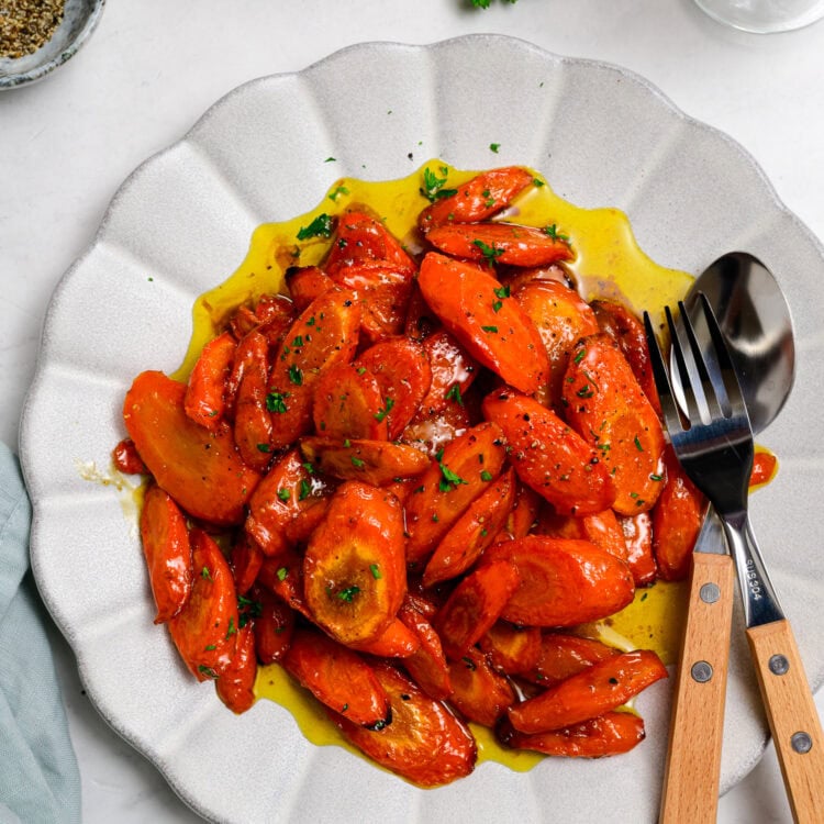 Sliced cooked Honey Garlic Carrots with herbs and a glossy sauce are served on a white plate, with serving utensils placed on the side.
