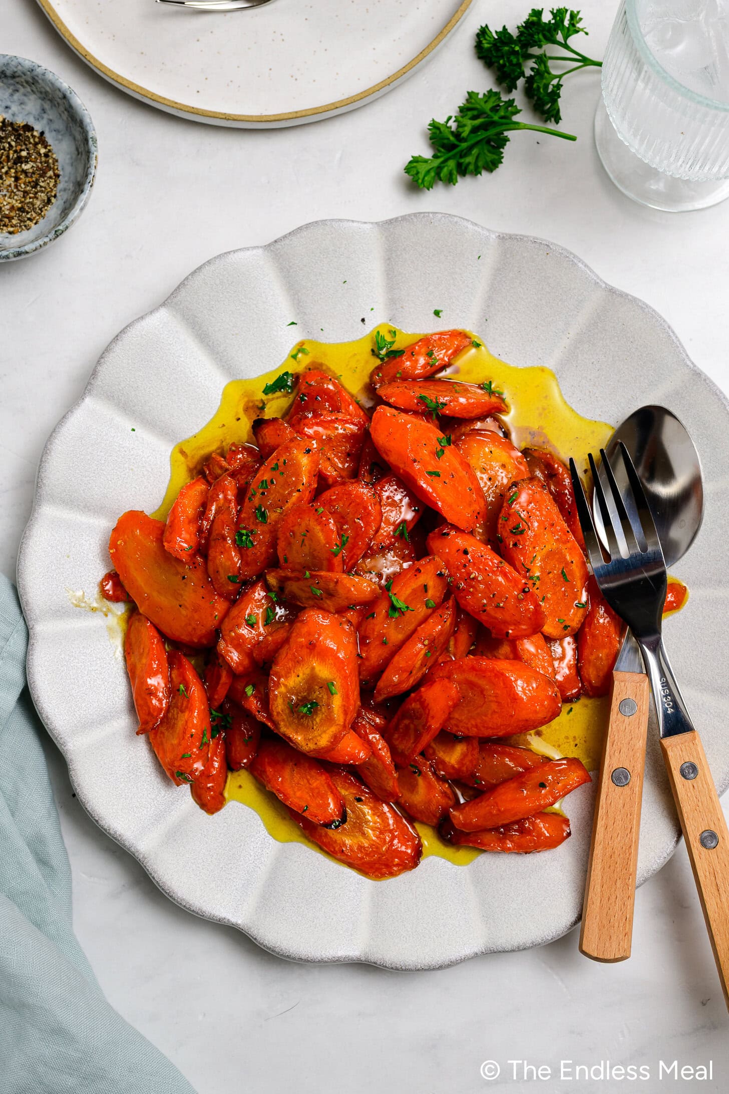 Sliced cooked Honey Garlic Carrots with herbs and a glossy sauce are served on a white plate, with serving utensils placed on the side.