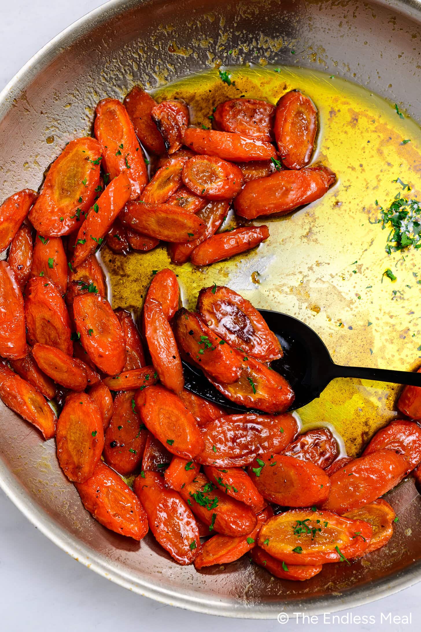Sliced honey garlic carrots in a pan with melted butter and herbs, with a black serving spoon resting among the carrots.