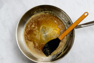 A stainless steel pan with browned butter and a black spatula resting inside on a white marble surface, perfect for preparing Honey Garlic Carrots.