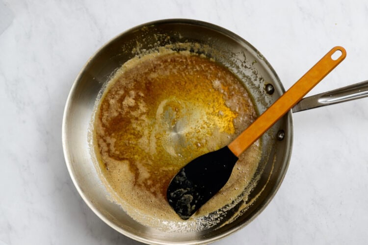 A stainless steel pan with browned butter and a black spatula resting inside on a white marble surface, perfect for preparing Honey Garlic Carrots.