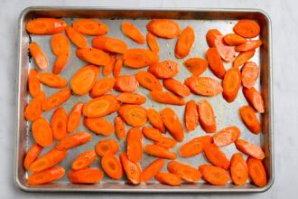 Sliced carrots coated with oil and seasoning are spread in a single layer on a metal baking sheet, ready to be transformed into delicious Honey Garlic Carrots.