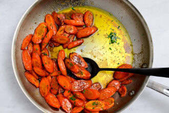 A skillet with sliced roasted Brown Butter Carrots glazed in a yellow honey garlic sauce, being stirred with a black spoon, on a light countertop.