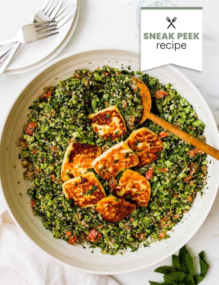 A bowl of tabbouleh salad topped with fried halloumi slices, served with a wooden spoon and garnished with fresh herbs.