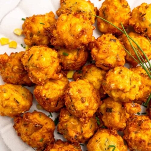 A plate of golden-brown corn fritters garnished with chives, the crispy corn fritters arranged in a cluster on a white plate.