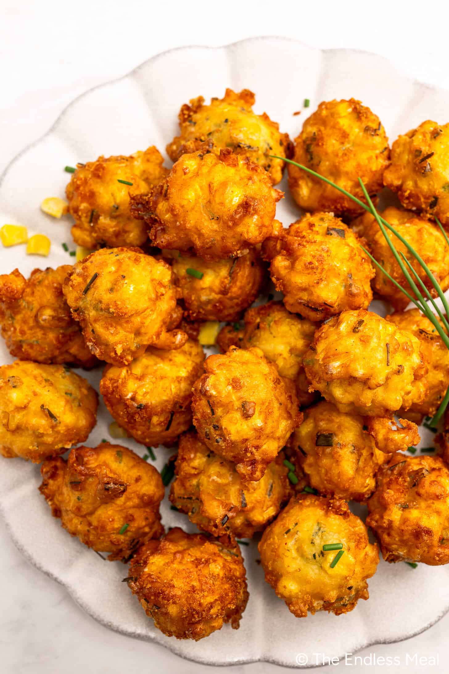 A plate of golden-brown corn fritters garnished with chives, the crispy corn fritters arranged in a cluster on a white plate.