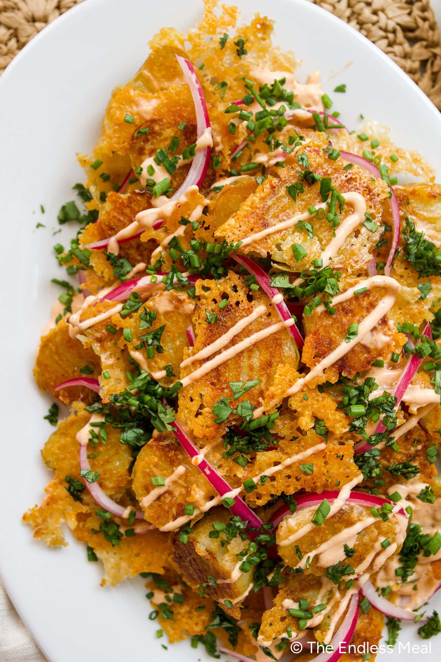 Crispy potatoes topped with melted cheese, sliced red onions, chopped herbs, and a drizzle of creamy sauce on a white plate.