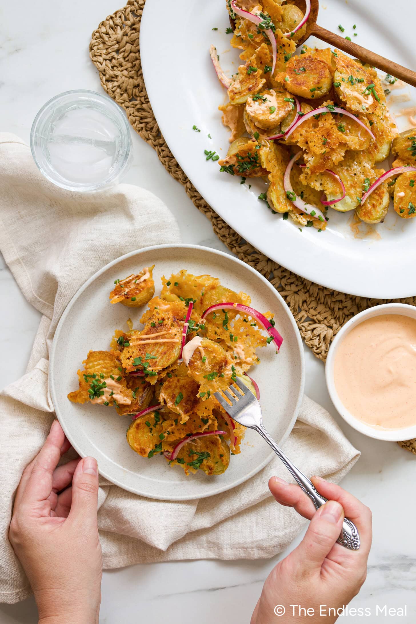 A hand holds a fork and a plate of crispy potato salad with herbs and red onions; a larger serving platter, glass, and napkin are also visible.