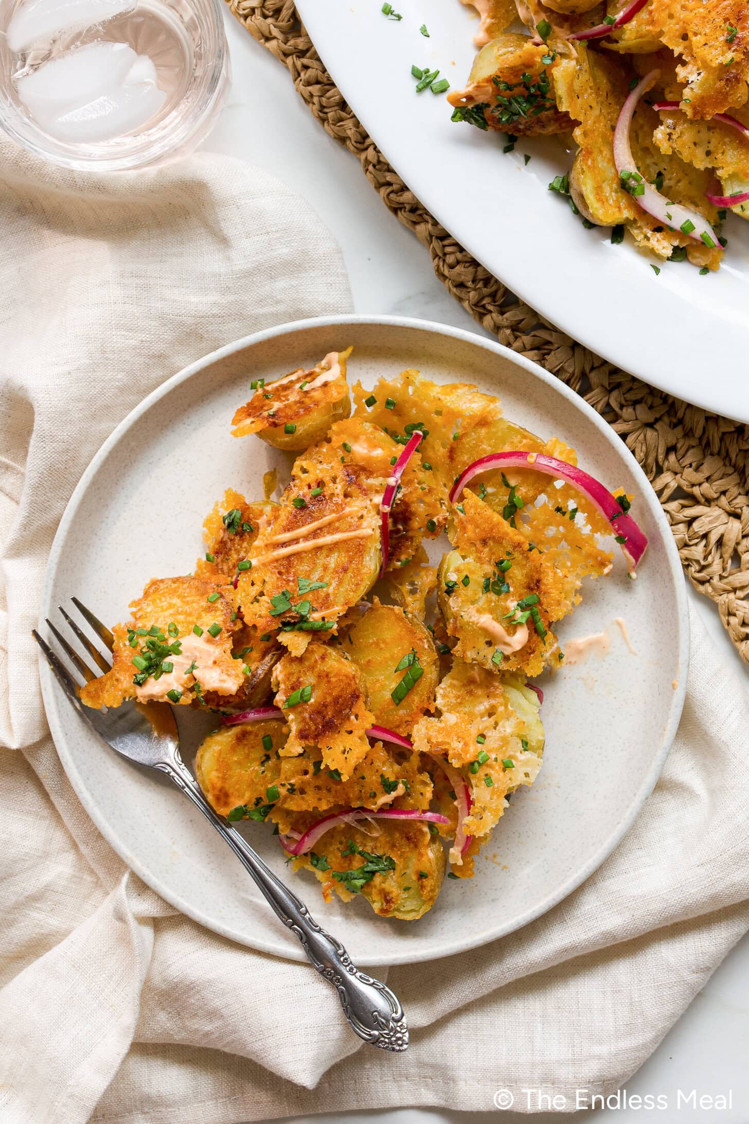 Plate of crispy smashed potatoes topped with melted cheese, red onion slices, and chopped chives, with a fork on the side and a glass of water in the background.