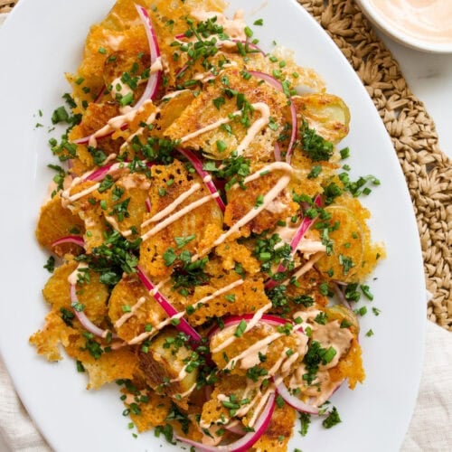Sliced roasted gochujang potatoes topped with melted cheese, creamy sauce, red onion slices, and chopped chives on a white oval plate.