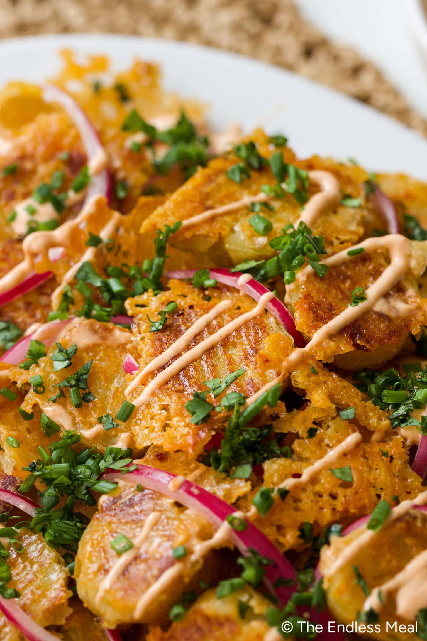 Close-up of crispy smashed potatoes topped with sliced red onions, chopped chives, and drizzled with a creamy orange sauce on a white plate.
