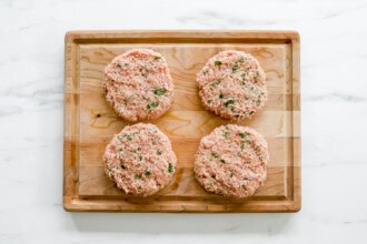 Image for recipe instruction: Four raw Teriyaki Chicken Burger patties with chopped herbs are arranged on a wooden cutting board atop a white marble surface.