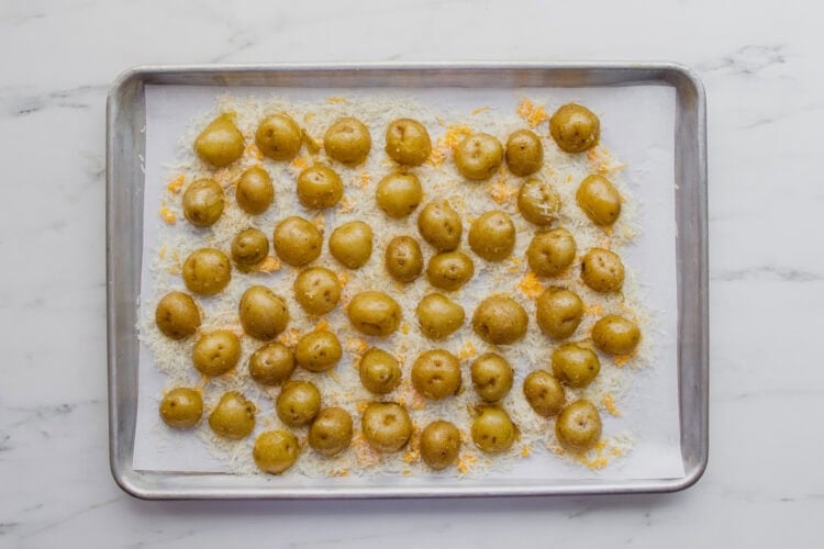 Sheet pan with halved baby potatoes on a layer of shredded cheese, ready for baking, on a white marble countertop.