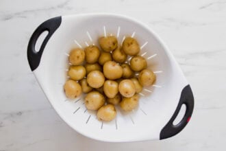 A white colander with red and black handles holds several small, whole potatoes on a white marble surface.