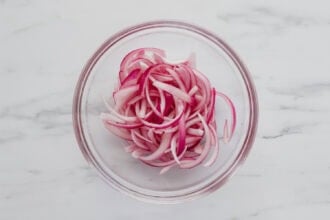 Thinly sliced red onions in a clear glass bowl on a white marble surface.