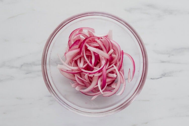Thinly sliced red onions in a clear glass bowl on a white marble surface.