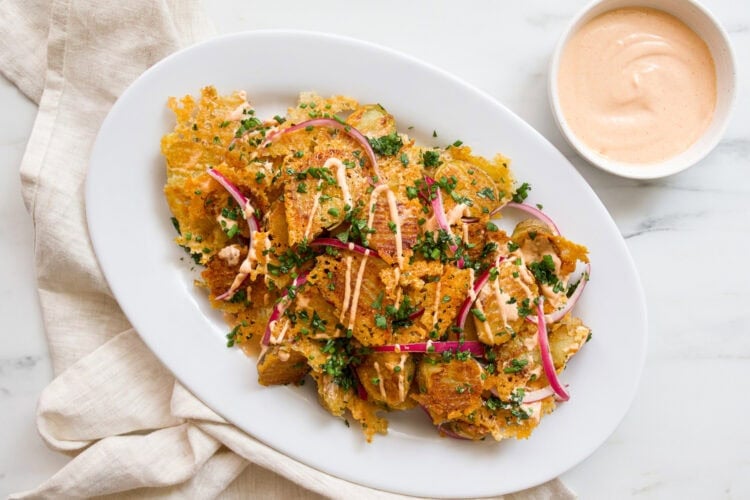 Oval plate with crispy smashed potatoes topped with cheese, red onions, herbs, and sauce; a bowl of creamy dipping sauce sits beside the plate on a marble surface.