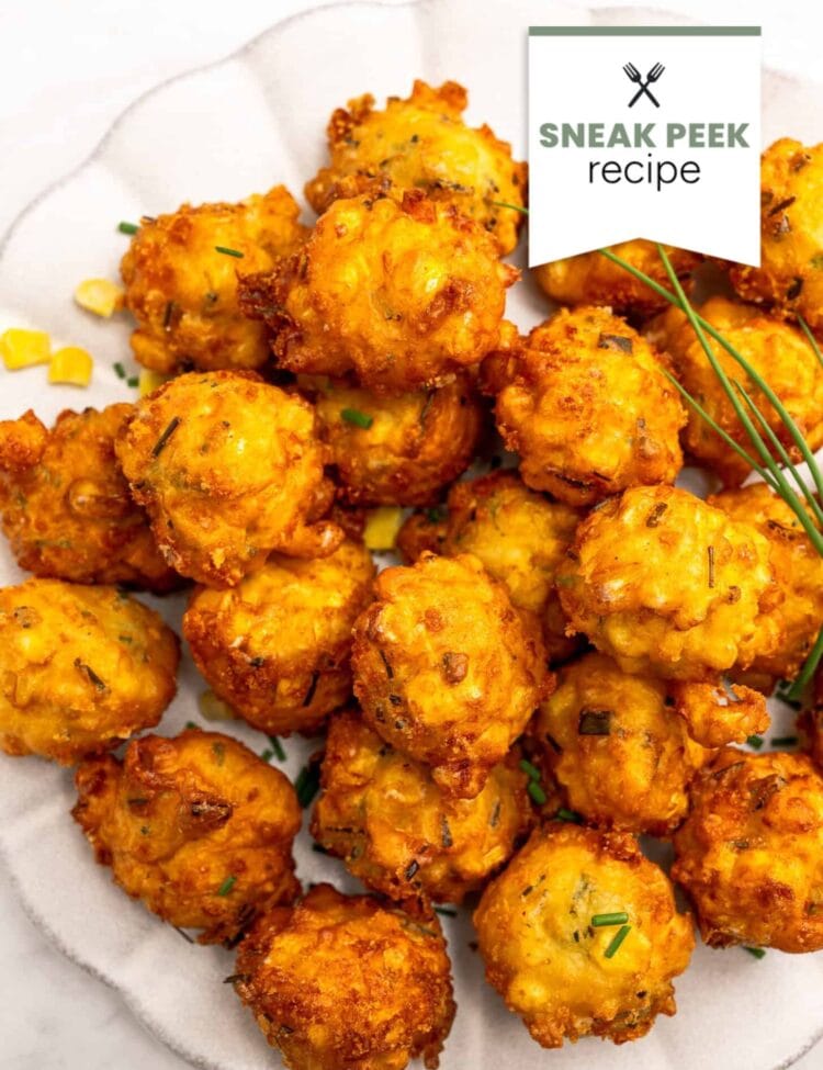 A plate of golden-brown corn fritters garnished with fresh chives and corn kernels, viewed from above. The Sneak Peek Recipe label in the upper right corner hints at a delicious new way to enjoy homemade corn fritters.