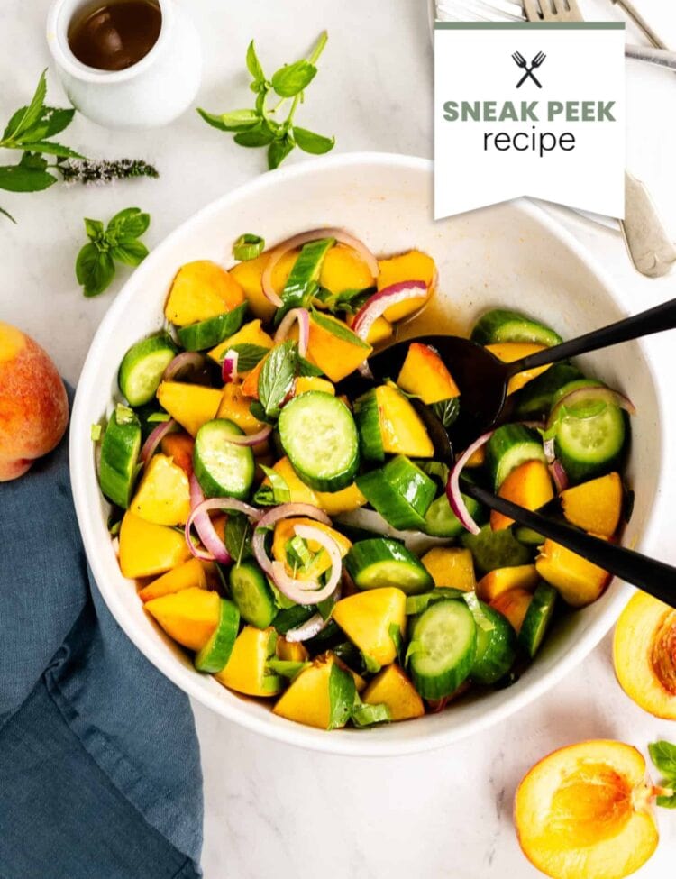 A bowl of Cucumber Peach Salad with red onion and herbs, served with tongs; peach halves and salad dressing rest nearby on a white surface.