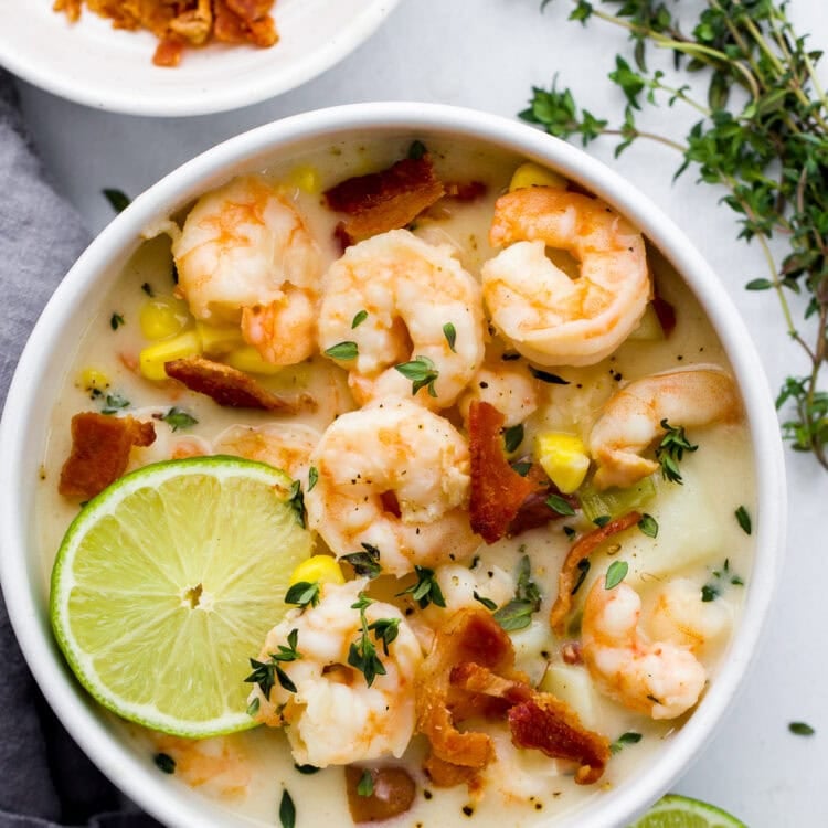 A bowl of creamy shrimp chowder garnished with bacon, fresh herbs, and lime slices, with extra bacon on a plate nearby—perfect for fans of creamy chowder.