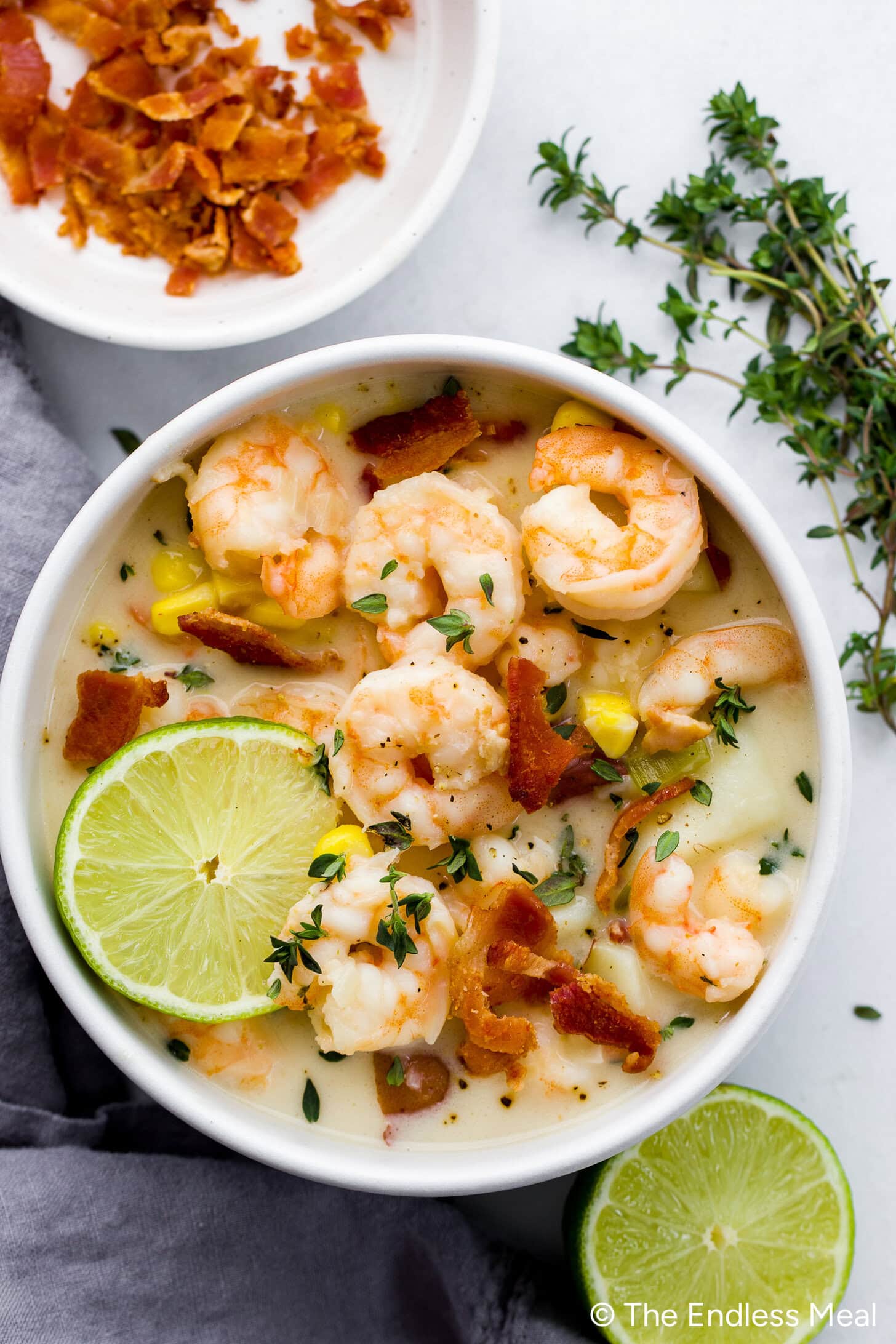 A bowl of creamy shrimp chowder garnished with bacon, fresh herbs, and lime slices, with extra bacon on a plate nearby—perfect for fans of creamy chowder.