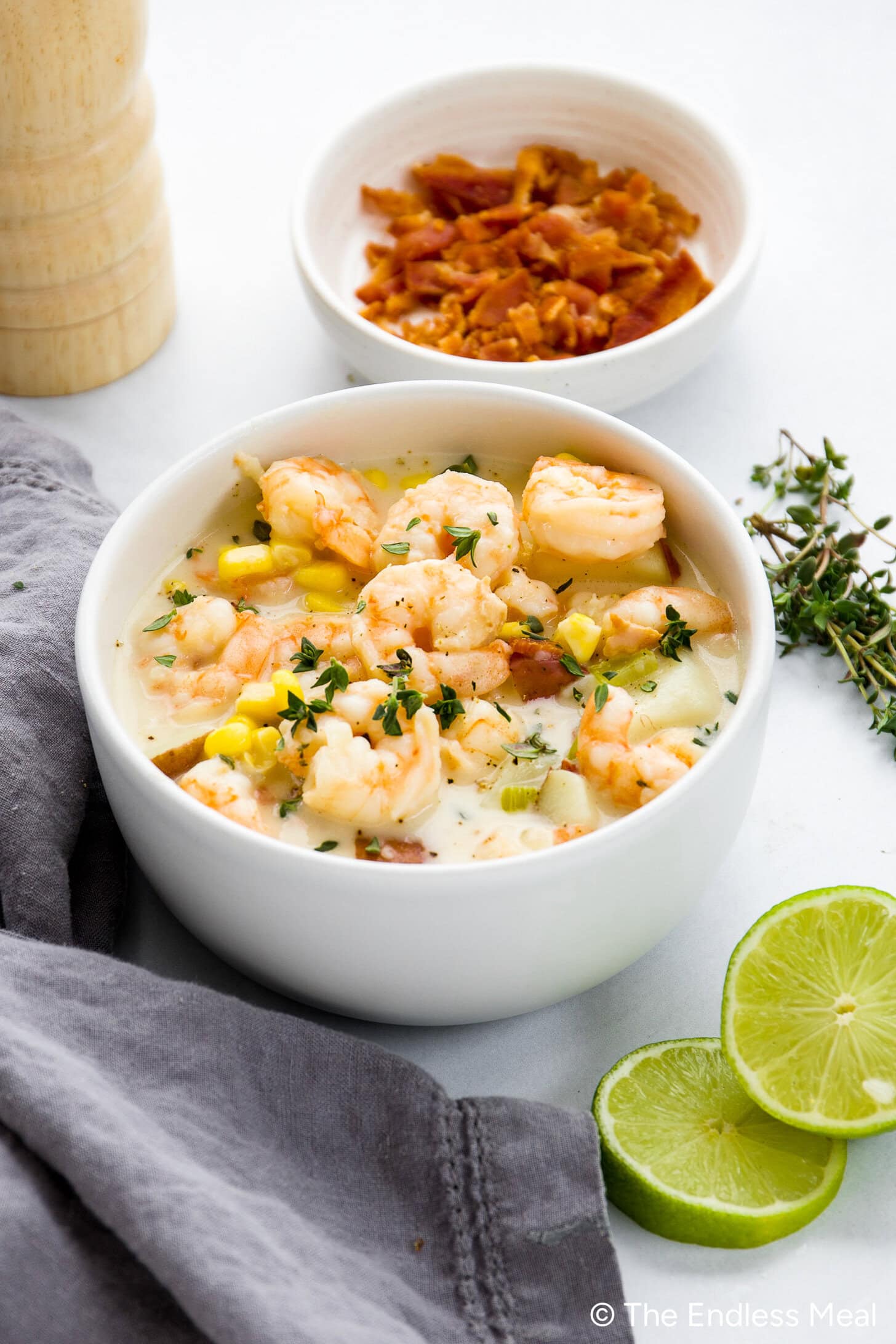 A white bowl filled with creamy corn and shrimp chowder, garnished with herbs, sits next to a bowl of bacon bits, lime slices, a gray napkin, and a wooden pepper mill.