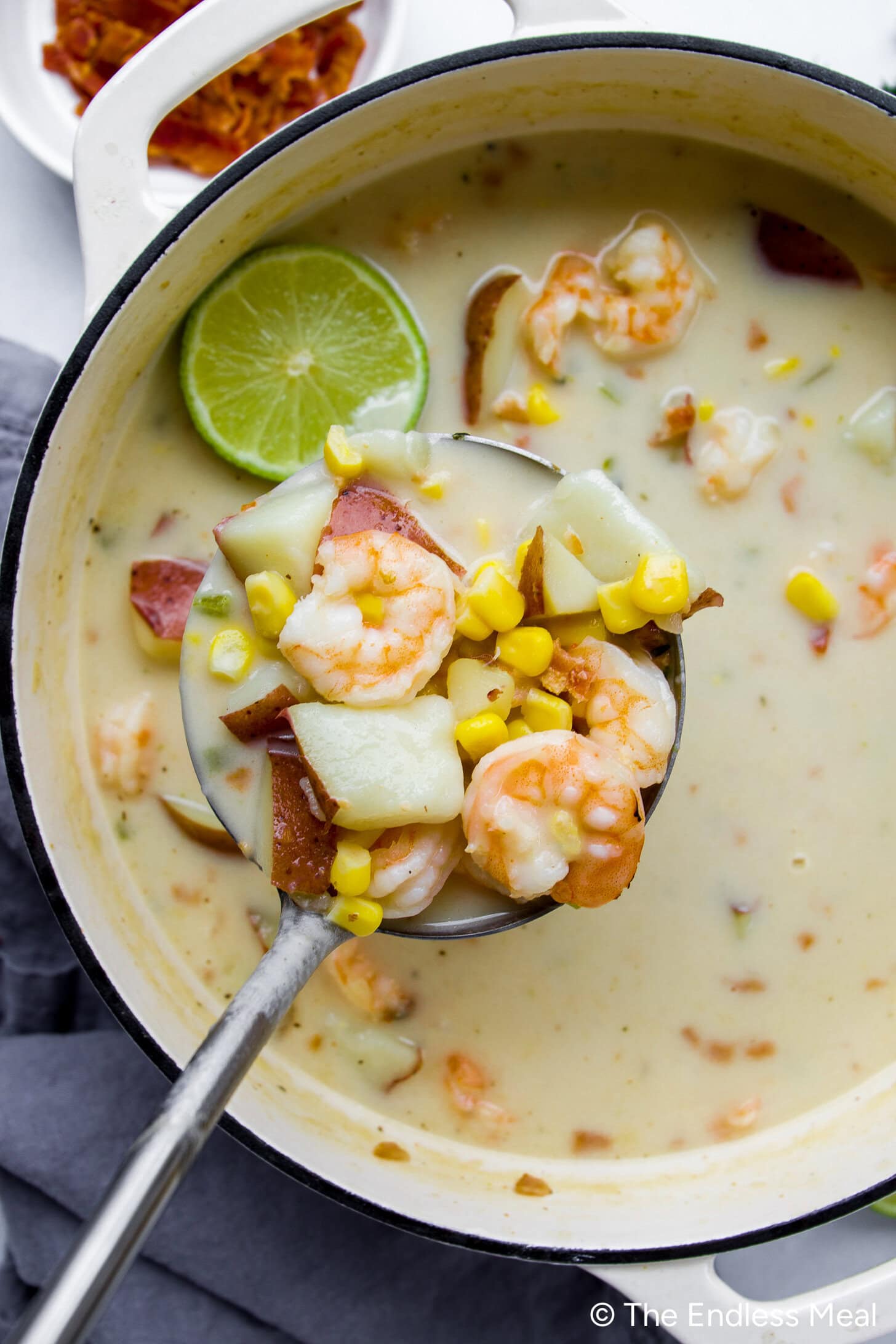 A ladle is lifting shrimp, corn, potatoes, and broth from a pot of creamy shrimp chowder, with a slice of lime floating on top.
