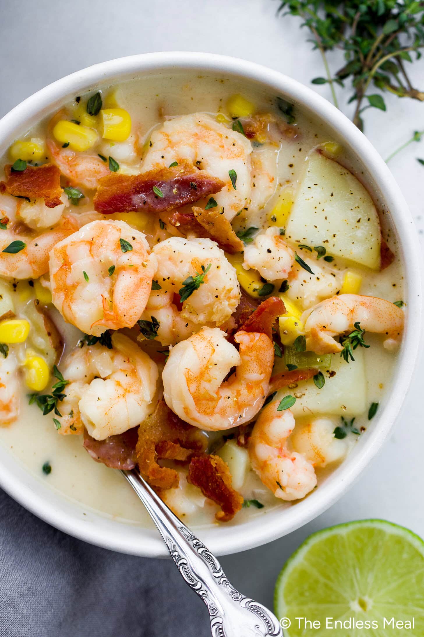 A bowl of creamy shrimp chowder with corn, bacon, potato chunks, and herbs, served with a spoon and a slice of lime nearby.